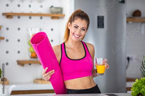 Happy shiny woman smiling after yoga practice, looking at camera. She's holding mat and juice. She's home in her kitchen. Portrait of a sporty smiling young woman, holding yoga mat in a roll