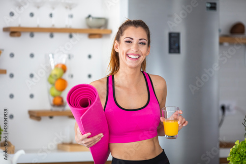Happy shiny woman smiling after yoga practice, looking at camera. She's holding mat and juice. She's home in her kitchen. Portrait of a sporty smiling young woman, holding yoga mat in a roll