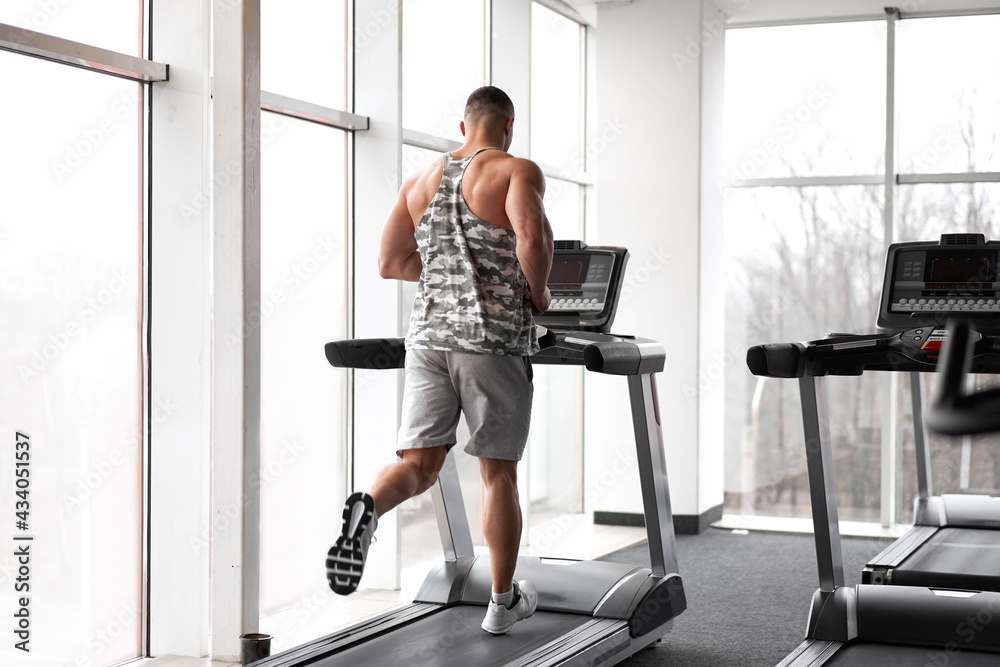 Fototapeta premium Muscular athletic bodybuilder fitness model running treadmill gym near big window