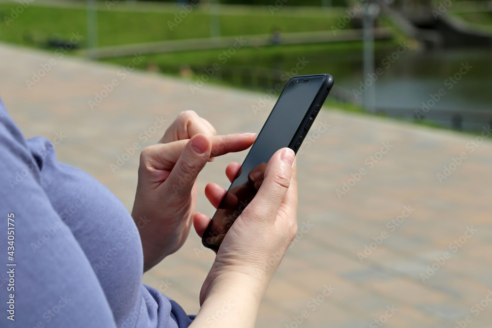 Woman with smartphone sitting in summer park. Close up of female hands with mobile phone on green nature background, concept of sms, online addiction, social media