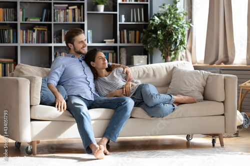 Happy young Caucasian couple renters or tenants relax on couch in living room look in distance dreaming thinking. Smiling dreamy man and woman rest on sofa at home imagine visualize together.