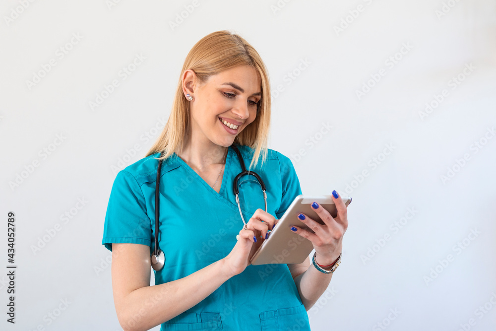 Young female clinician doctor in scrubs using touchpad while communicating with patients online. Portrait Of Smiling Female Doctor Wearing Scrubs In Hospital Corridor Holding Digital Tablet Young female clinician doctor in scrubs using touchpad while communicating with patients online. Portrait Of Smiling Female Doctor Wearing Scrubs In Hospital Corridor Holding Digital Tablet