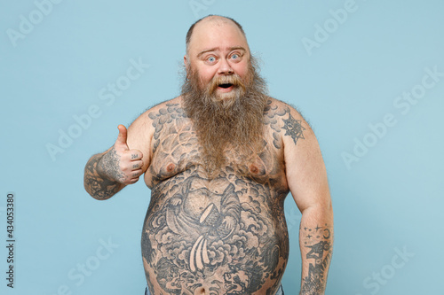 Young happy fat pudge obese chubby overweight blue-eyed bearded man 30s has big belly with naked tattooed torso showing thumb up isolated on pastel blue background studio. People lifestyle concept photo