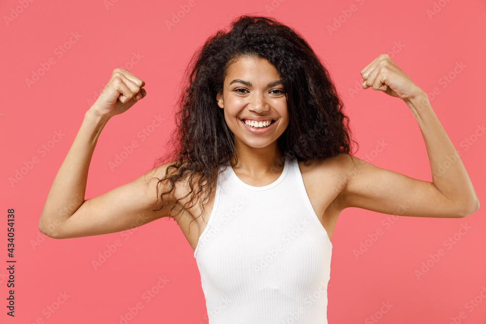Young fun strong sporty fitness african american woman 2s wearing casual white tank shirt showing biceps muscles on hand demonstrating strength power isolated on pink background. Lifestyle concept