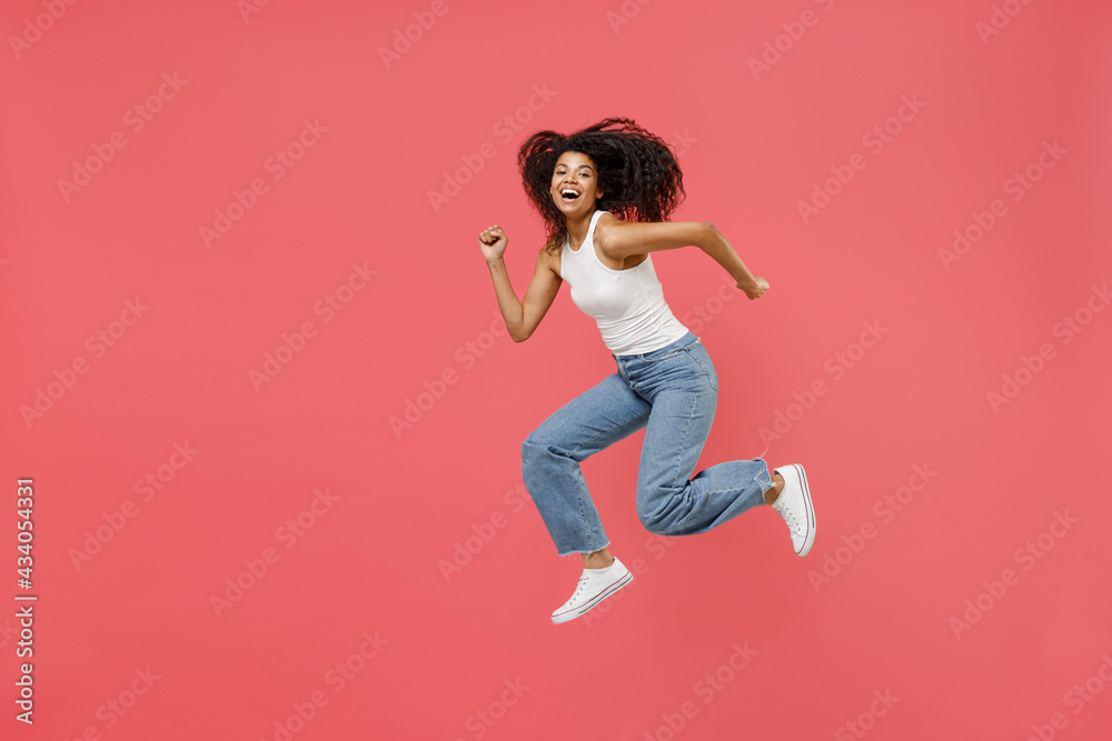 Full length side view young fun happy positive cheerful energetic african american woman in casual white tank shirt jump high run fast hurrying up isolated on pink color background studio portrait
