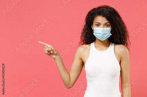 Young african woman in white tank shirt sterile face mask ppe coronavirus virus covid-19 flu on lockdown quarantine point index finger aside on workspace area copy space isolated on pink background