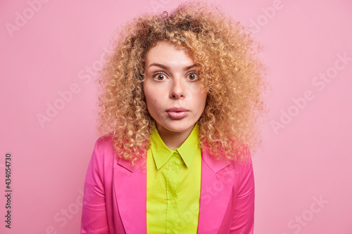Impressed speechless curly haired young woman in bright formal clothes cannot believe her eyes being amazed by something unexpected isolated over pink background. Human face expressions concept