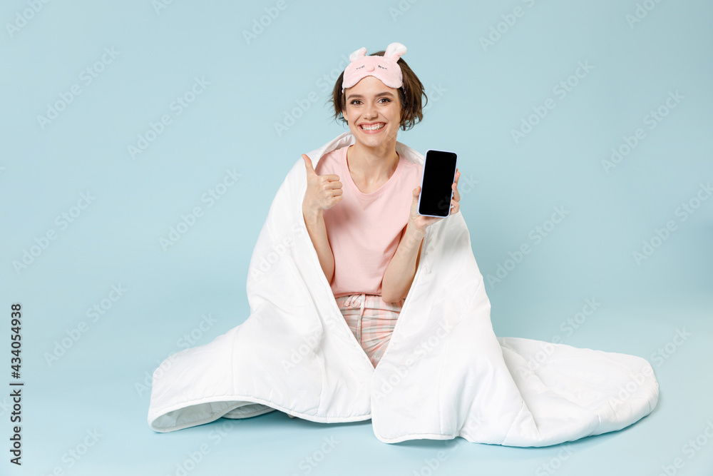 Naklejka premium Young woman in pajamas jam sleep mask rest at home sit wrap cover blanket duvet use mobile phone worskspace area show like gesture isolated on pastel blue background Good mood night bedtime concept.
