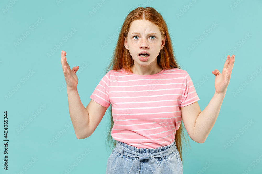 Little indignant angry expressive confused redhead kid girl 12-13 years old in pink striped t-shirt spread hands arms isolated on pastel blue background studio Children lifestyle childhood concept