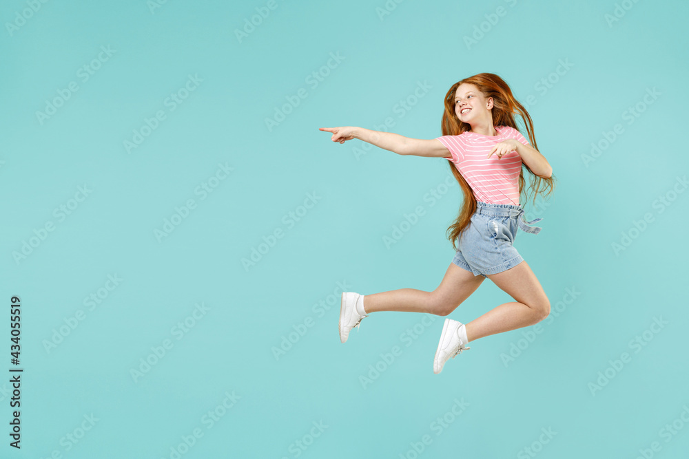 Full length little redhead kid girl 12-13 year old in pink striped t-shirt jump point index finger aside on copy space mock up isolated on pastel blue background Children lifestyle childhood concept.