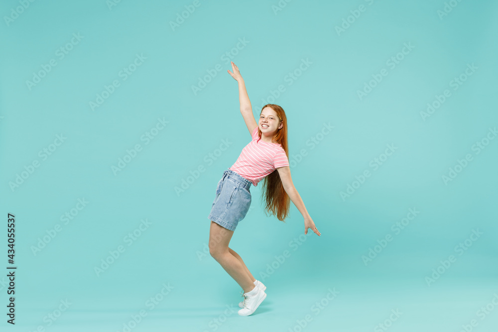 Full length little redhead kid girl 12-13 years old in pink striped t-shirt stand on toes leaning back dance isolated on pastel blue background studio portrait. Children lifestyle childhood concept.