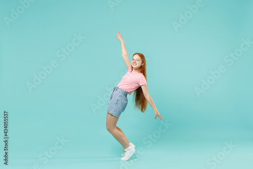 Fototapeta Naklejka Na Ścianę i Meble -  Full length little redhead kid girl 12-13 years old in pink striped t-shirt stand on toes leaning back dance isolated on pastel blue background studio portrait. Children lifestyle childhood concept.