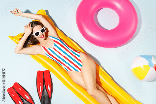 Top view young sexy woman slim body wear striped one-piece swimsuit lies on inflatable mattress ring ball hotel pool isolated on pastel blue color background. Summer vacation sea rest sun tan concept.