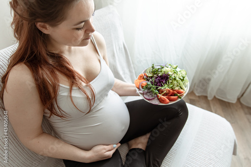 Pregnant woman with a plate of fresh vegetable salad at home on the couch.