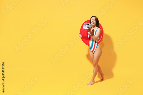 Full length happy sexy woman slim body wear red blue swimsuit hold inflatable rubber ring scream megaphone isolated on vivid yellow color background studio. Summer hotel pool sea rest sun tan concept