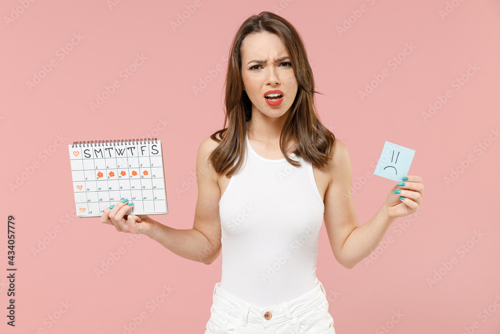 Young indignant woman in white clothes hold in hand female periods calendar check menstruation days card sign sad smiley isolated on pastel pink background. Medical healthcare gynecological concept. Young indignant woman in white clothes hold in hand female periods calendar check menstruation days card sign sad smiley isolated on pastel pink background. Medical healthcare gynecological concept.