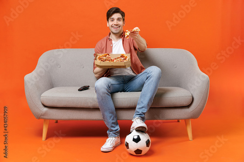 Fototapeta Naklejka Na Ścianę i Meble -  Young man football fan in shirt support favorite team with soccer ball sit on sofa at home watch tv live stream give pizza slice isolated on orange background People sport leisure lifestyle concept