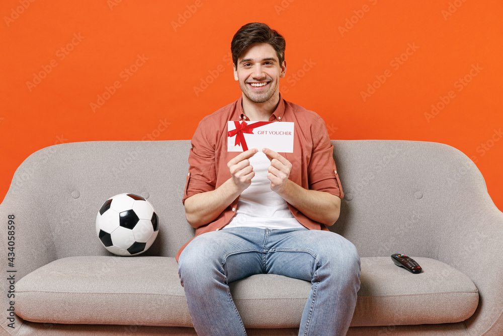 Young fun man football fan wearing shirt support team with soccer ball  sitting on sofa home