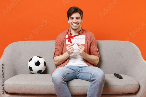 Young fun man football fan wearing shirt support team with soccer ball sitting on sofa home watching tv live stream hold gift voucher flyer mock up isolated on orange background. People sport concept