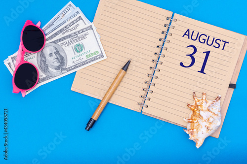 Travel concept flat lay - notepad with the date of 31 august pen, glasses, dollars and seashell on blue background. photo