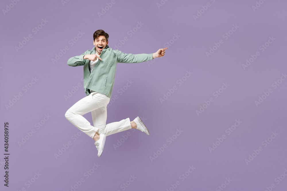 Full length fun excited cool young man in casual mint shirt white t-shirt jump high point index finger aside on workspace area mock up isolated on purple violet background. People lifestyle concept