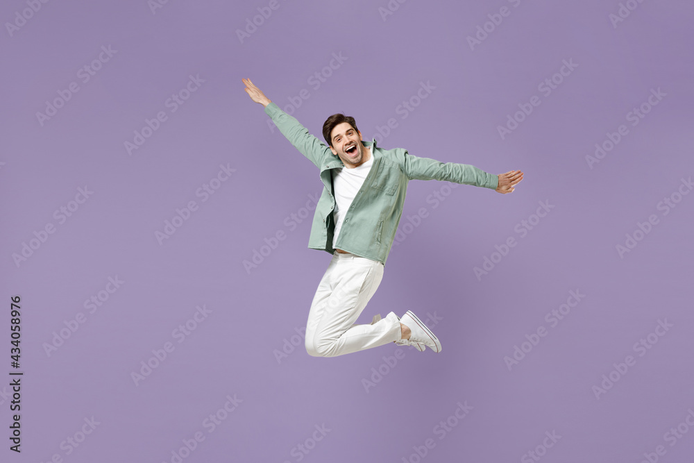 Fototapeta premium Full length excited overjoyed energetic cool young man in casual mint shirt white t-shirt jump high with outstretched hands look camera isolated on purple violet background. People lifestyle concept