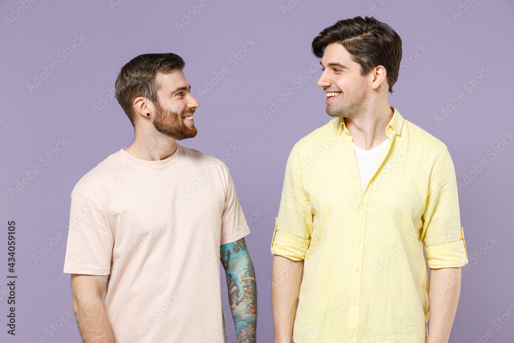 Two young smiling cheerful caucasian positive happy cool men friends together wearing casual t-shirt looking to each other isolated on purple color background studio portrait People lifestyle concept Two young smiling cheerful caucasian positive happy cool men friends together wearing casual t-shirt looking to each other isolated on purple color background studio portrait People lifestyle concept
