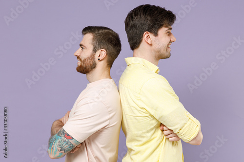 Side view of two young happy men friends together in casual t-shirt stand back to back hold hands crossed folded look to each other isolated on purple background People lifestyle friendship concept. © ViDi Studio
