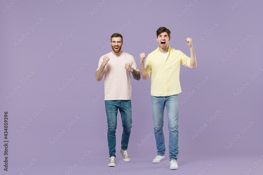 Full length overjoyed two young happy men friends together in casual t-shirt walking do winner gesture clench fist isolated on purple background studio portrait People friendship lifestyle concept Full length overjoyed two young happy men friends together in casual t-shirt walking do winner gesture clench fist isolated on purple background studio portrait People friendship lifestyle concept