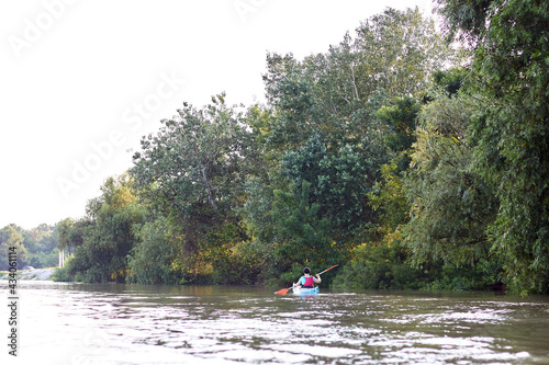 Woman rowing in blue kayak near shoreline with green trees at Danube river at summer