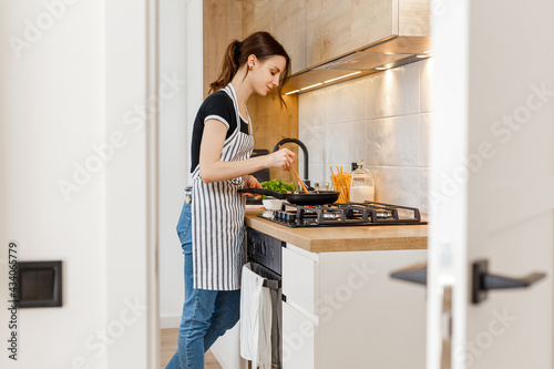 Young woman in apron cooking healthy food at modern home kitchen. Preparing meal with frying pan on gas stove. Concept of domestic lifestyle, happy housewife leisure and culinary hobby