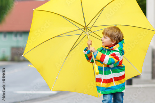Little toddler boy playing with big yellow umbrella on rainy day. Happy positive child running through rain and puddles. Kid with rain clothes and rubber boots. Children activity on bad weather day.