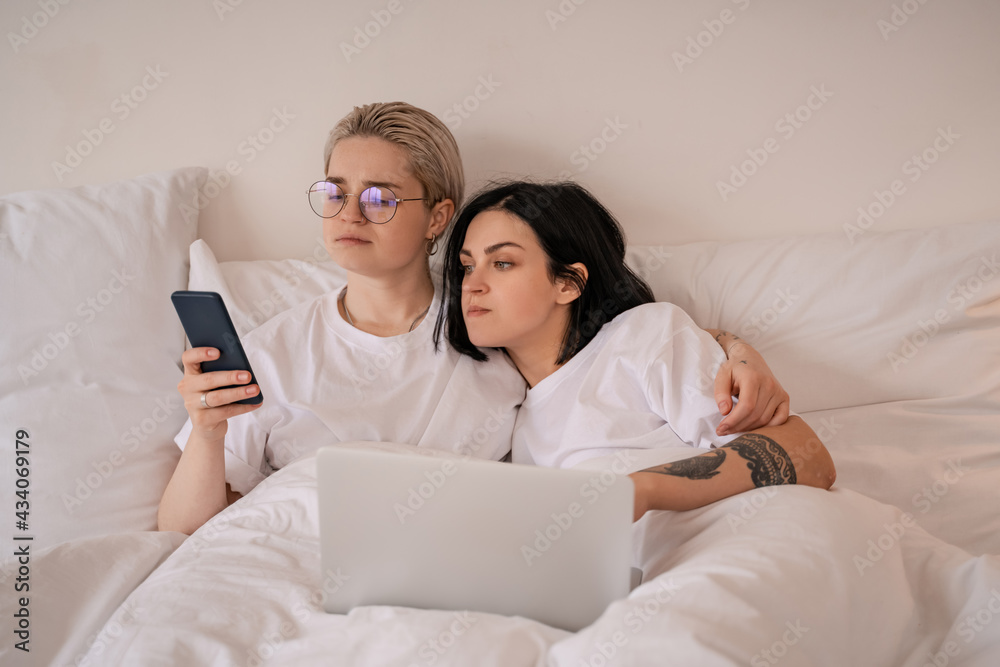 young lesbian couple lying in bed and looking at smartphone near laptop young lesbian couple lying in bed and looking at smartphone near laptop
