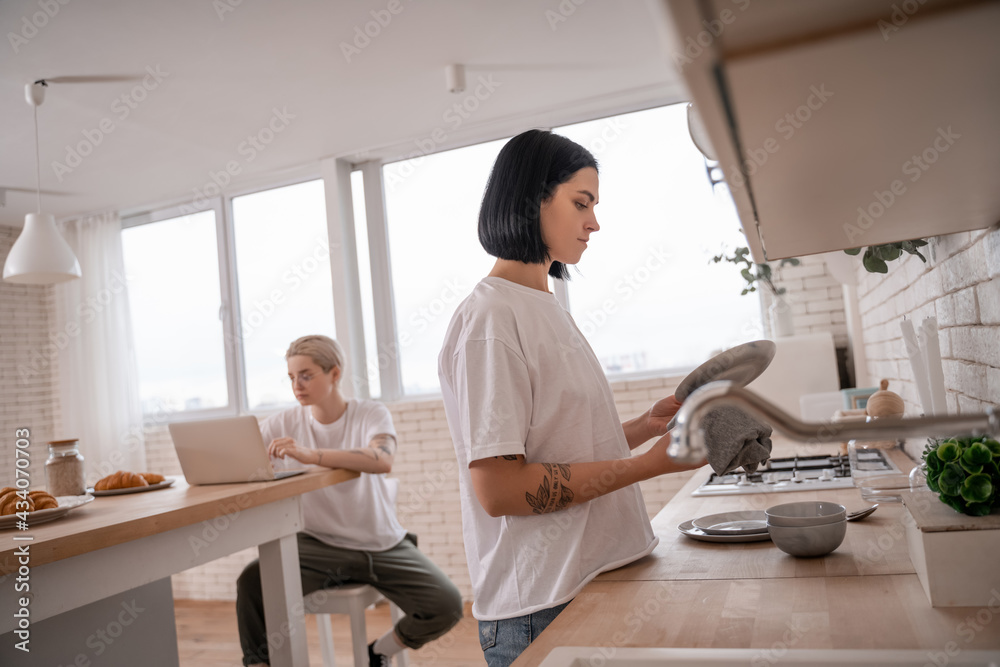 Obraz premium brunette woman washing plate near girlfriend using laptop on blurred background