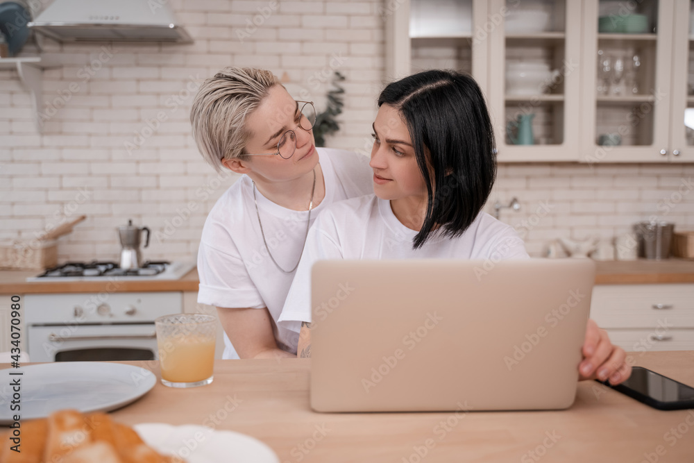 young woman looking at girlfriend using laptop in kitchen young woman looking at girlfriend using laptop in kitchen
