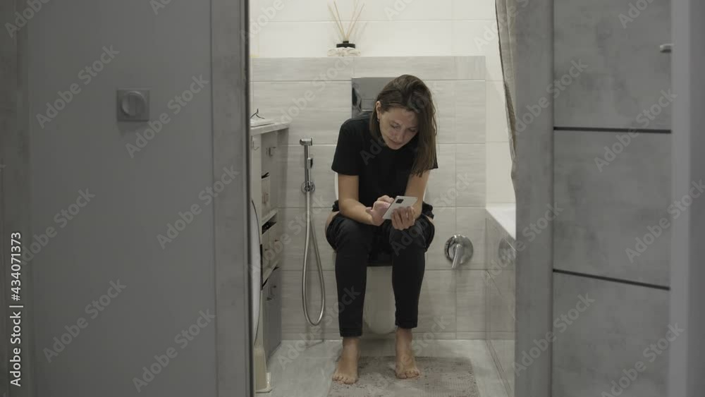 Young woman in casual clothes in bathroom, sitting on toilet bowl and using cellphone. Woman On The Toilet Looking At Her Mobile Phone