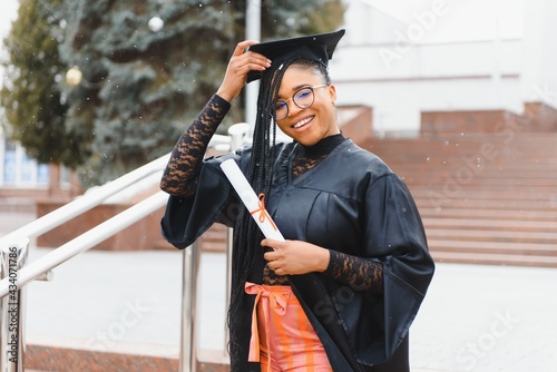 pretty african college student in graduation cap and gown in front of school building