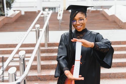 pretty african college student in graduation cap and gown in front of school building