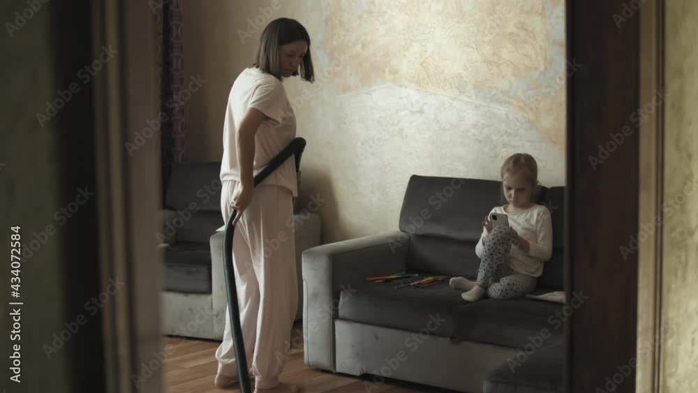 A young woman in pajamas vacuuming carpet with vacuum cleaner in the living room. Little girl sitting on couch and watch TV during her mother cleaning up the apartment A young woman in pajamas vacuuming carpet with vacuum cleaner in the living room. Little girl sitting on couch and watch TV during her mother cleaning up the apartment