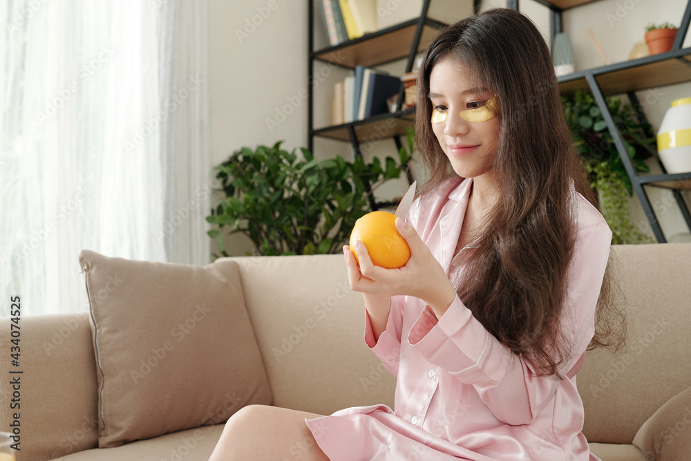 Happy young pretty woman peeling fresh orange for breakfast when sitting on sofa in pajamas after waking up Happy young pretty woman peeling fresh orange for breakfast when sitting on sofa in pajamas after waking up