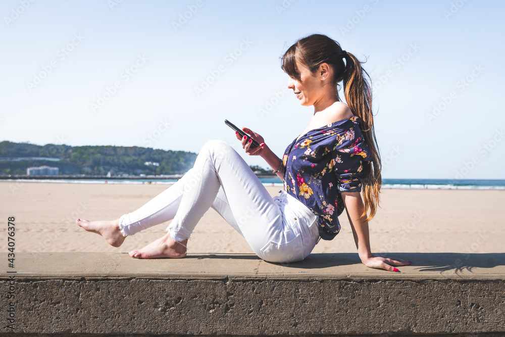 Young caucasian woman using a mobile phone sitting on the side of a beach promenade, at Hendaia -Basque Country.