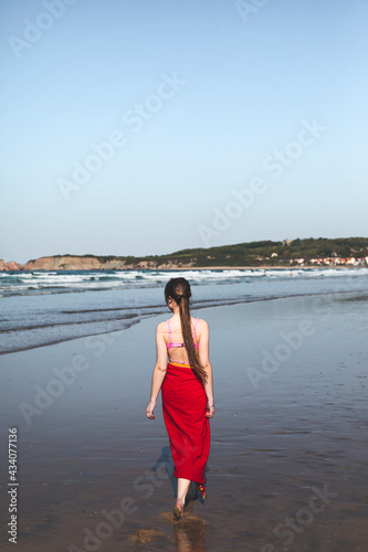 Young woman wearing a red towel and a pink bikini walking at the water edge on the beach