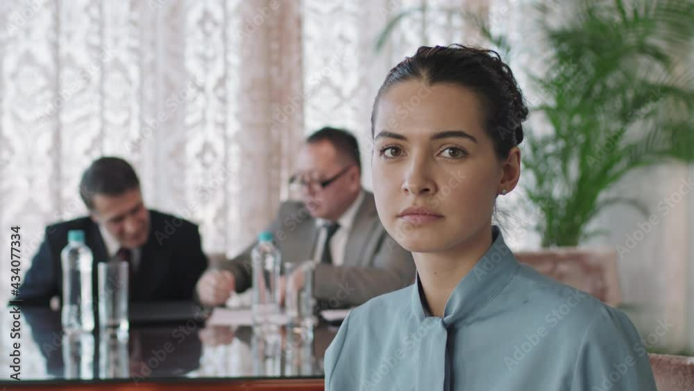 Portrait of young businesswoman looking at camera sitting at conference table in meeting room while business partners signing contract in background. Suitable for deepfake generation or face swap Portrait of young businesswoman looking at camera sitting at conference table in meeting room while business partners signing contract in background. Suitable for deepfake generation or face swap
