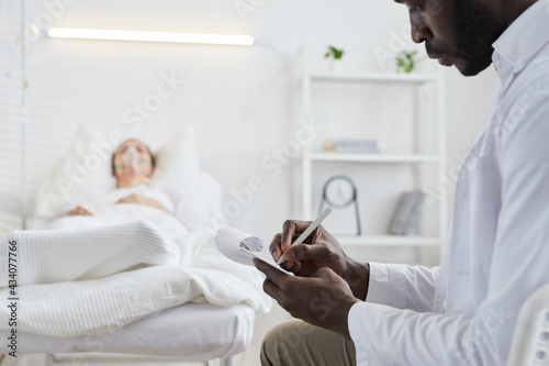Close-up of African doctor writing prescription in medical card for patient who lying on hospital bed