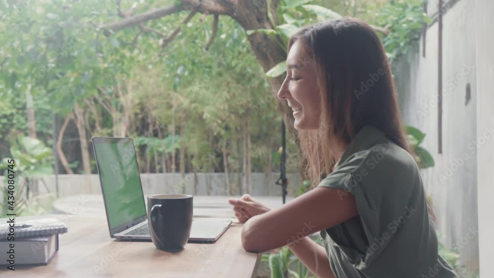 Side view waist-up of young Caucasian woman sitting by desk in foreground of green tropical terrace on summer day, having video call on laptop with green screen, looking cheerful