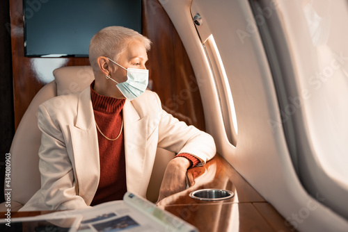 Businesswoman in medical mask looking at plane window near magazine and digital tablet