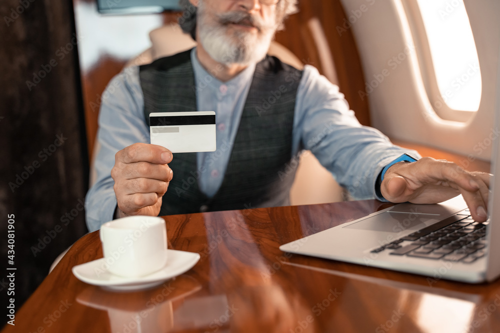 Fototapeta premium Cropped view of credit card in hand of businessman using laptop near cup in plane