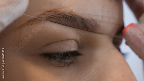 Stylist's hands working with special thread to mark new contour of brows closeup. Professional styling, correction and shaping procedures of female eyebrows in beauty salon