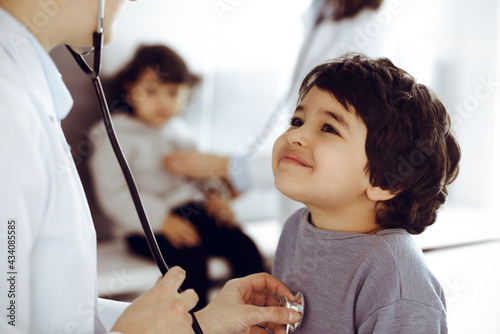 Doctor-woman examining a child patient by stethoscope. Cute arab boy and his brother at physician appointment. Medicine concept