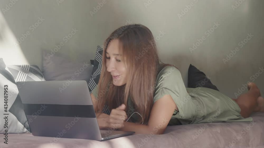 Full shot of happy Caucasian woman wearing wired headphones, lying on stomach on sofa with decorative pillows, talking, using laptop computer for online call Full shot of happy Caucasian woman wearing wired headphones, lying on stomach on sofa with decorative pillows, talking, using laptop computer for online call